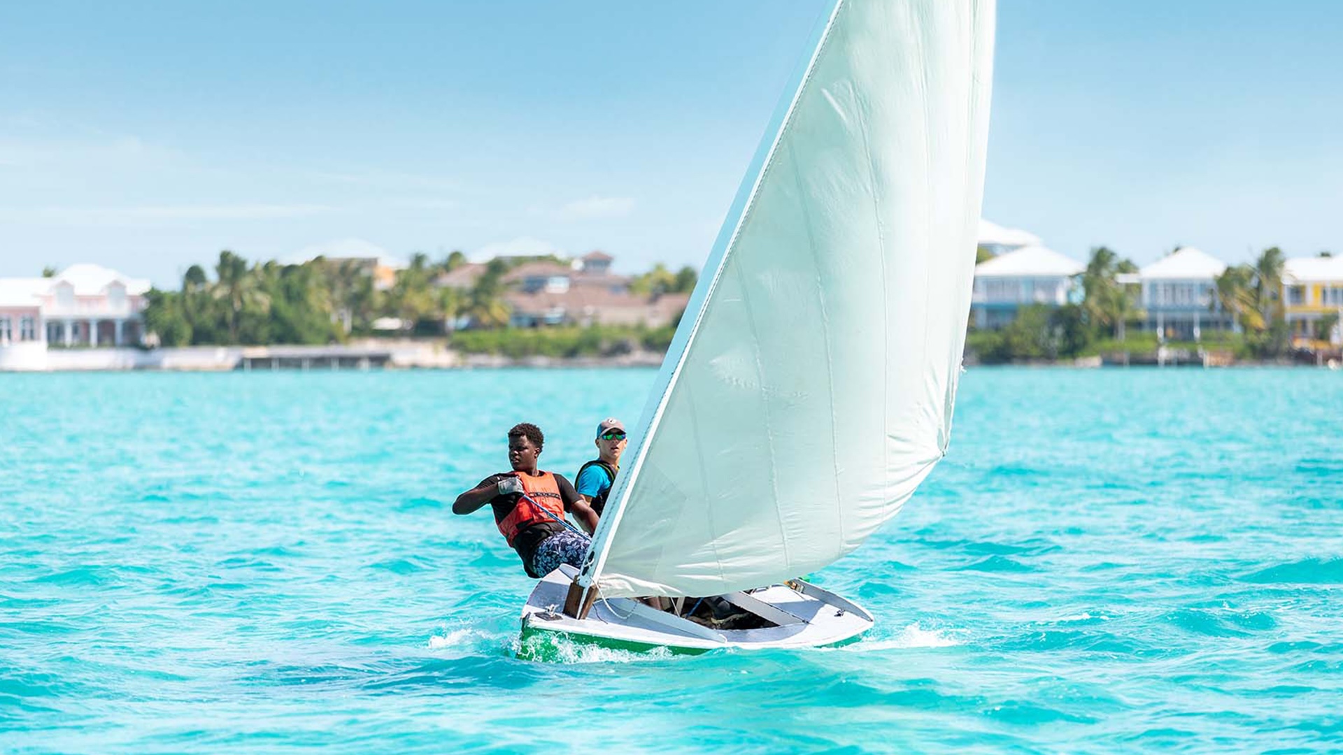 Dos jóvenes navegando un dinghy a vela en agua turquesa con costa de palmeras