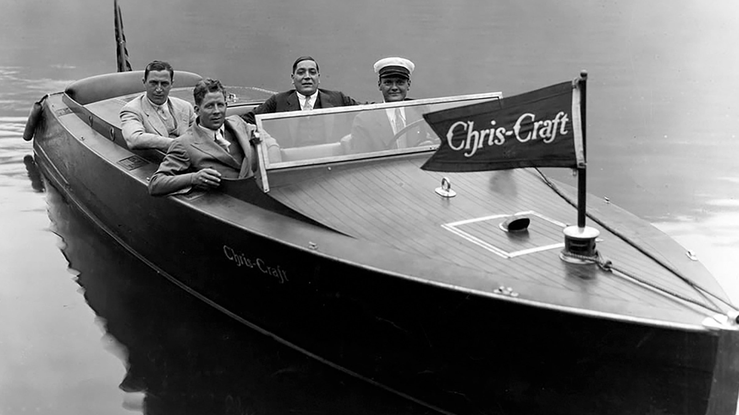 Four men in jackets and ties, one in a captain's cap, seated aboard a classic Chris-Craft, period photograph